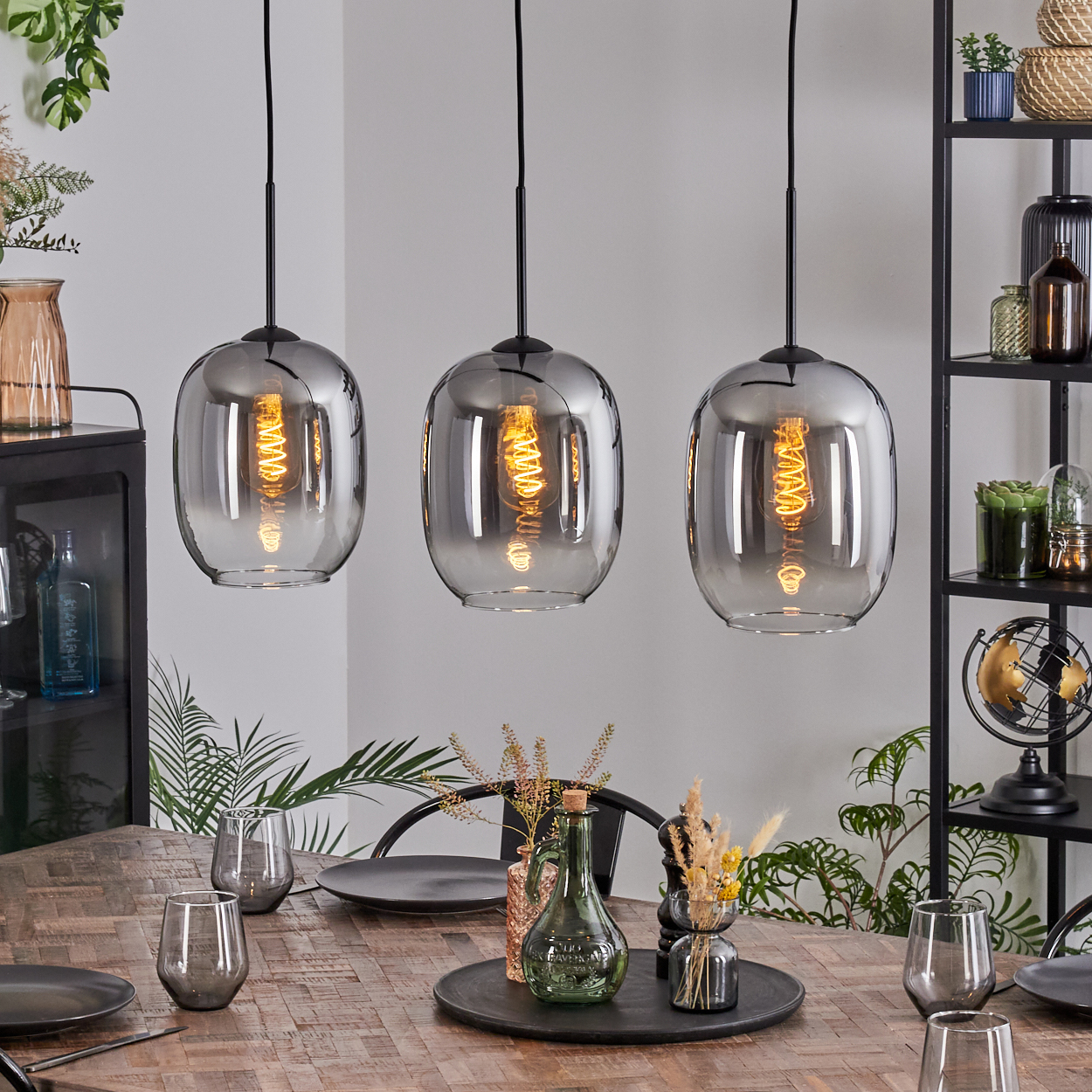 Drei Pendelleuchten hängen über einem Holztisch, der mit Tellern, Gläsern und einem Mittelstück gedeckt ist. Regale und Dekorationen sind im Hintergrund.