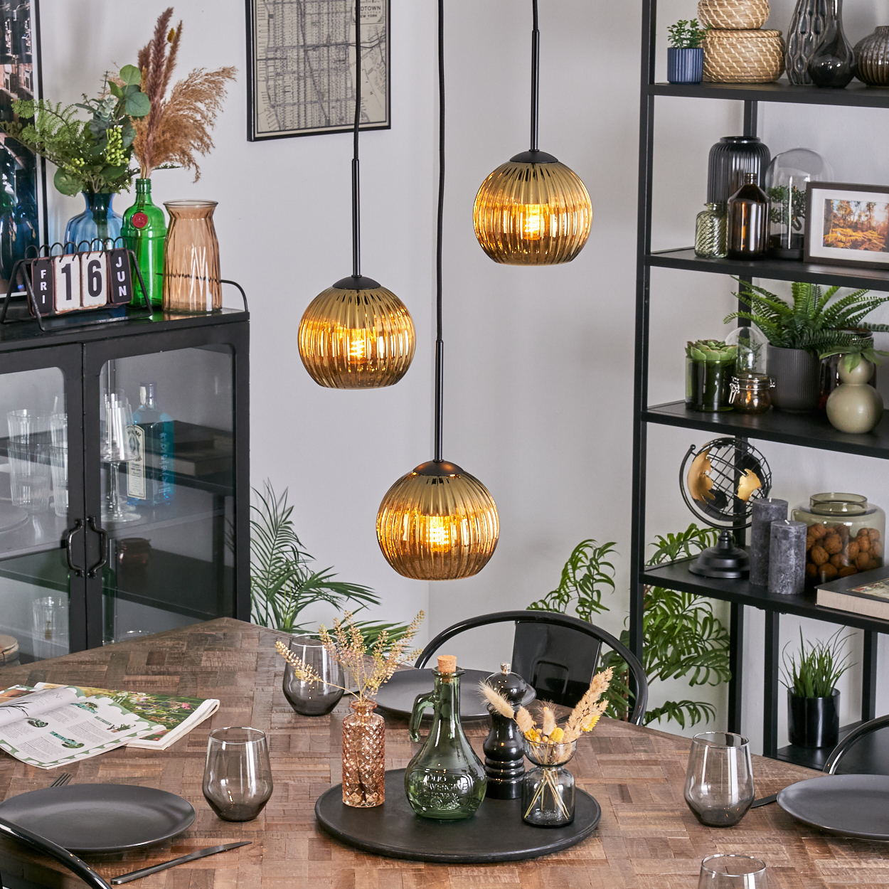 Esszimmer mit Holztisch, Hängelampen und verschiedenen Dekorationen. Enthält Teller, Gläser und ein schwarzes Metallregal.