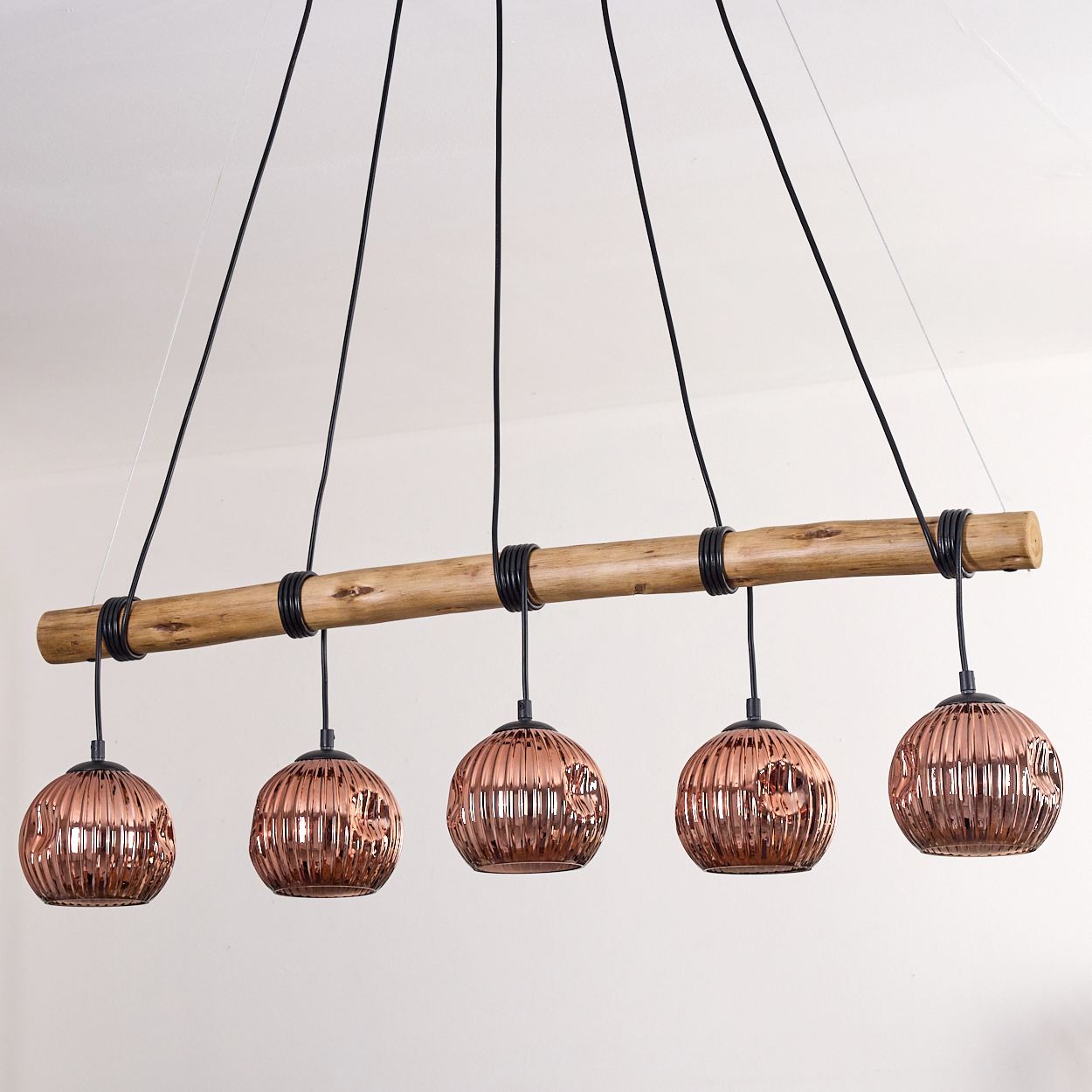 Moderne Leuchte mit fünf kupferfarbenen, gerippten Glasschirmen, die an einem Holzbalken hängen. Schwarze Kabel und ein weißer Hintergrund.