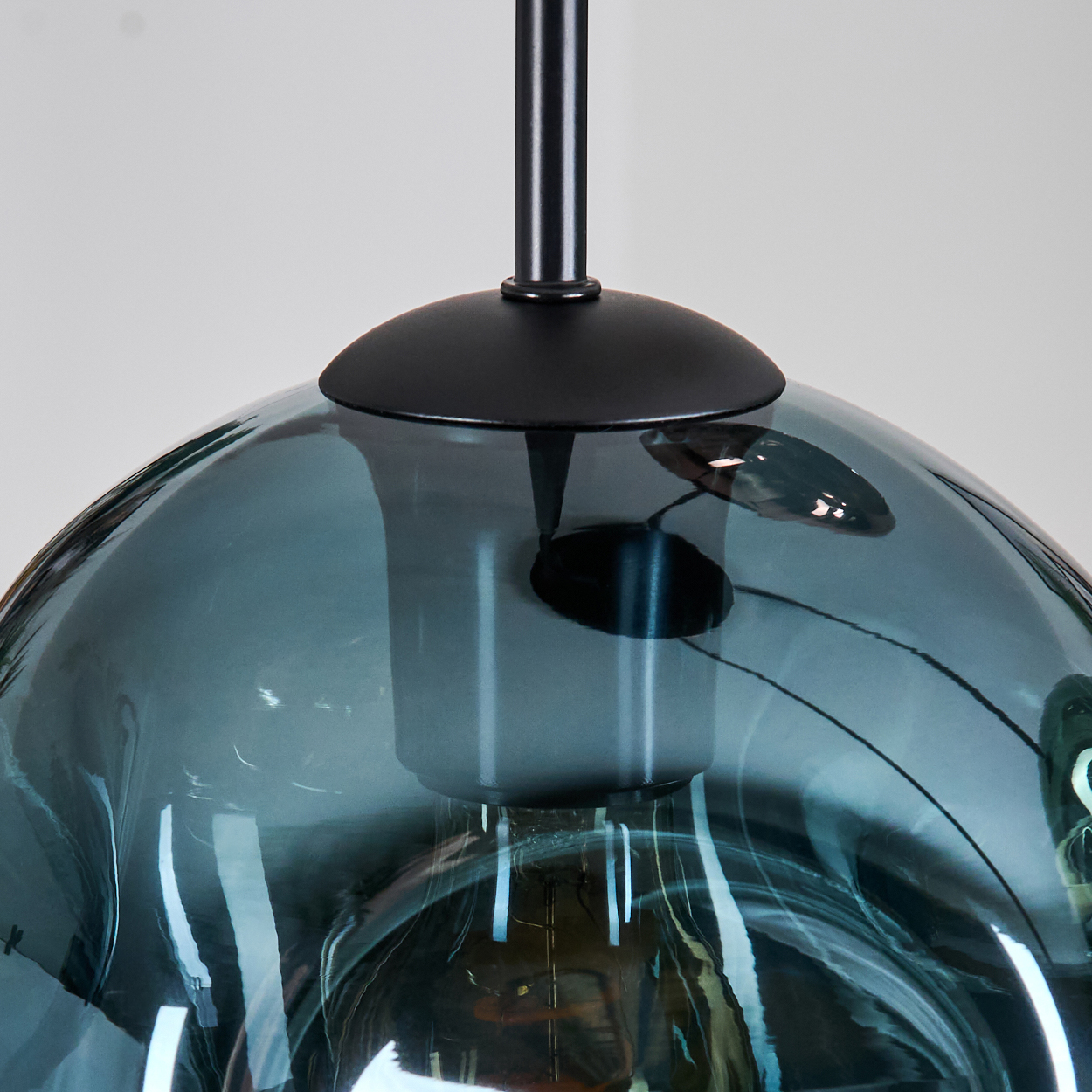 Nahaufnahme einer tealfarbenen Glas-Pendelleuchte mit schwarzem Metallschaft und Baldachin, vor weißem Hintergrund.
