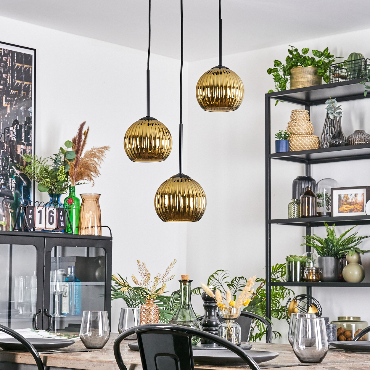 Esszimmer mit goldenen Pendelleuchten. Tisch mit Gläsern und Dekor. Regale mit Pflanzen und Dekor im Hintergrund.