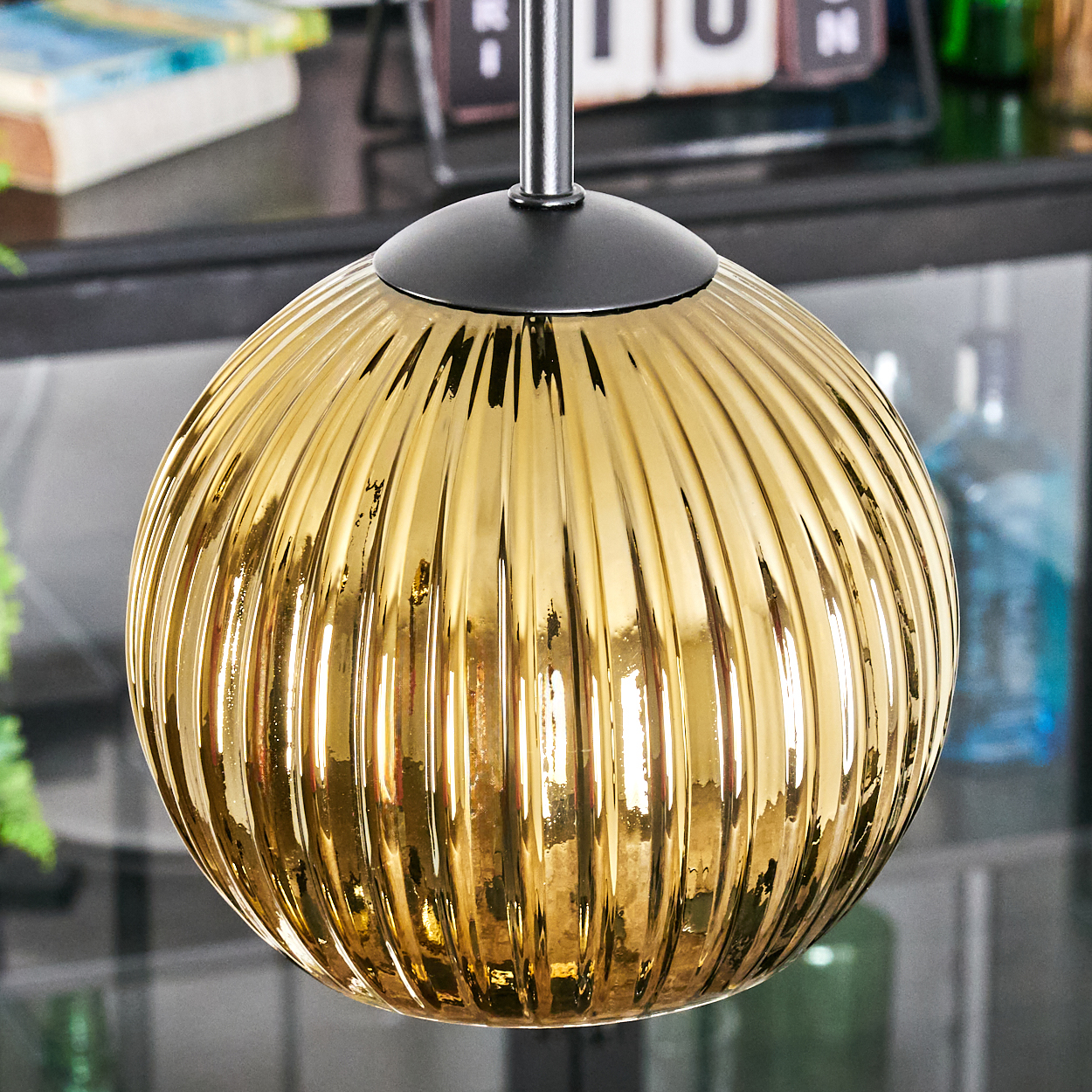Eine goldene, gerippte Glas-Pendelleuchte hängt. Sie hat eine schwarze Kappe und eine Metallstange. Im Hintergrund verschwommene Objekte und ein Kalender.