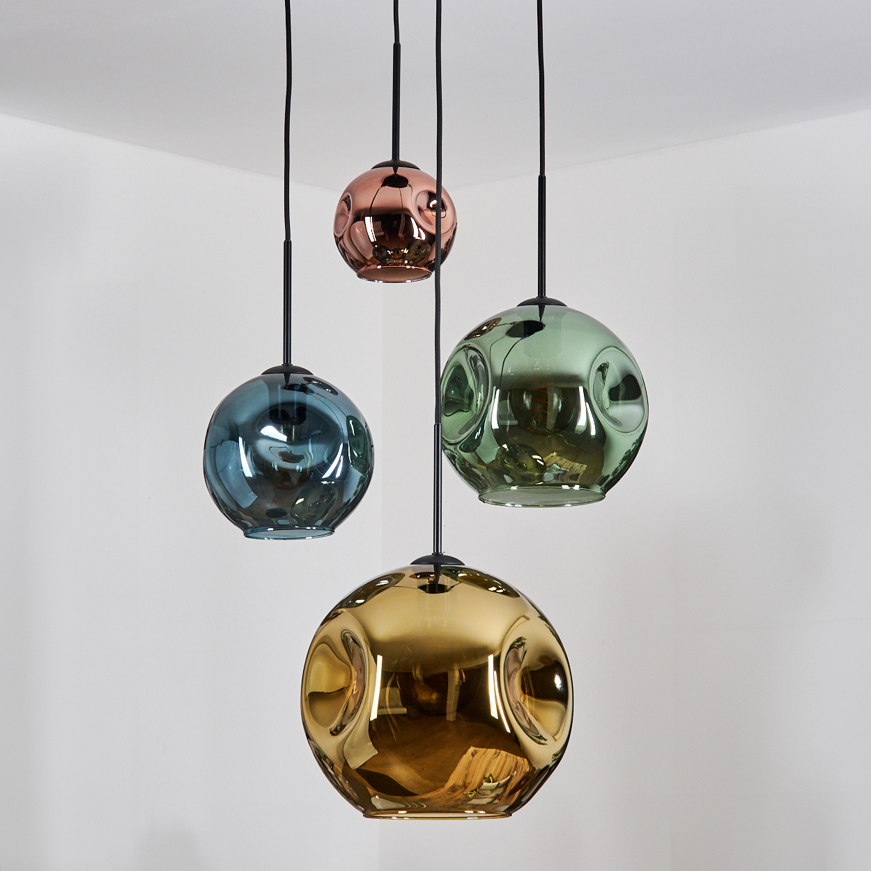 Vier kugelförmige Pendelleuchten in verschiedenen Farben: Blau, Grün, Kupfer und Gold. Sie hängen an schwarzen Kabeln vor einer weißen Wand.