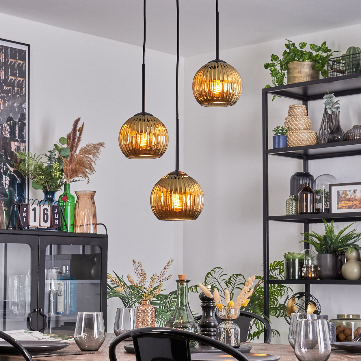 Esszimmer mit drei goldenen Pendelleuchten. Tisch mit Gläsern und Dekor. Schwarzes Regal mit Pflanzen und Dekorationsartikeln.