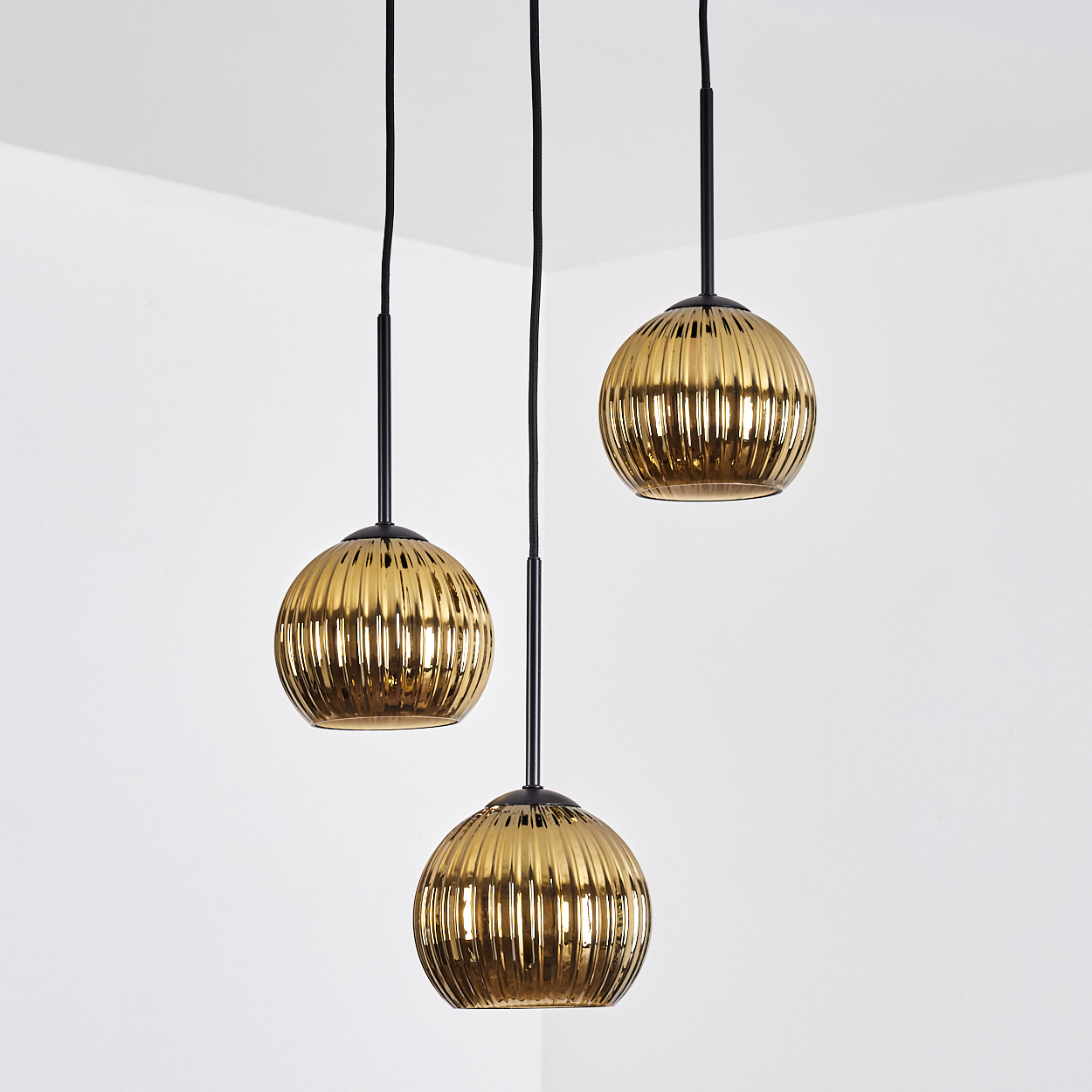 Drei goldene, geriffelte Glas-Pendelleuchten hängen an schwarzen Kabeln vor einem weißen Eckhintergrund.