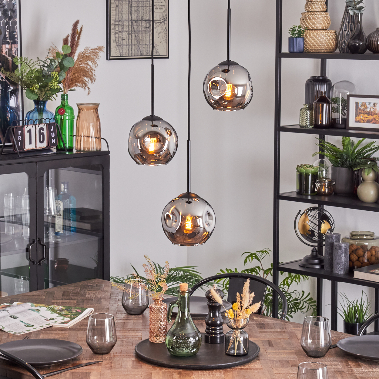 Esszimmer mit Holztisch, Hängelampen, Regalen mit Dekor und einem schwarzen Schrank. Der Tisch hat Teller und Gläser.