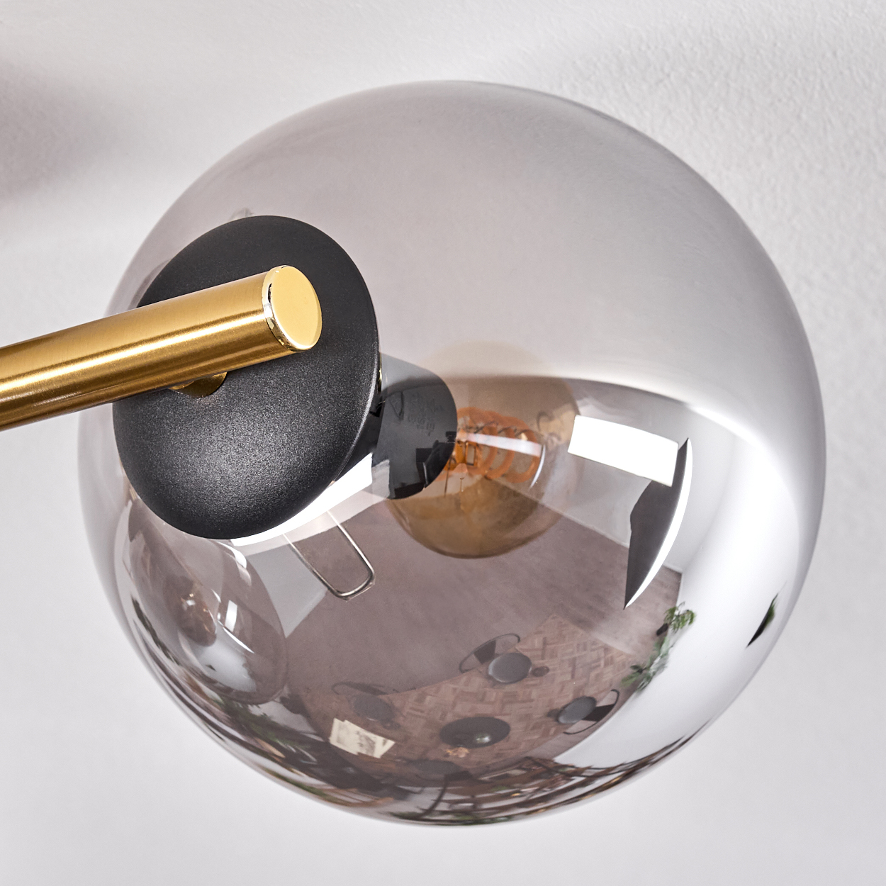 Eine moderne Lampe mit kugelförmigem, grauem Glasschirm. Ein goldener Arm und ein schwarzer Sockel tragen den Schirm. Im Inneren spiegelt sich ein Raum.