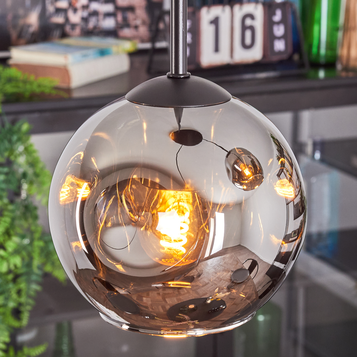Eine runde, rauchfarbene Glashängelampe mit einer leuchtenden Wendelbirne im Inneren. Reflektierende Oberfläche. Hintergrund: Kalender und Pflanzen.