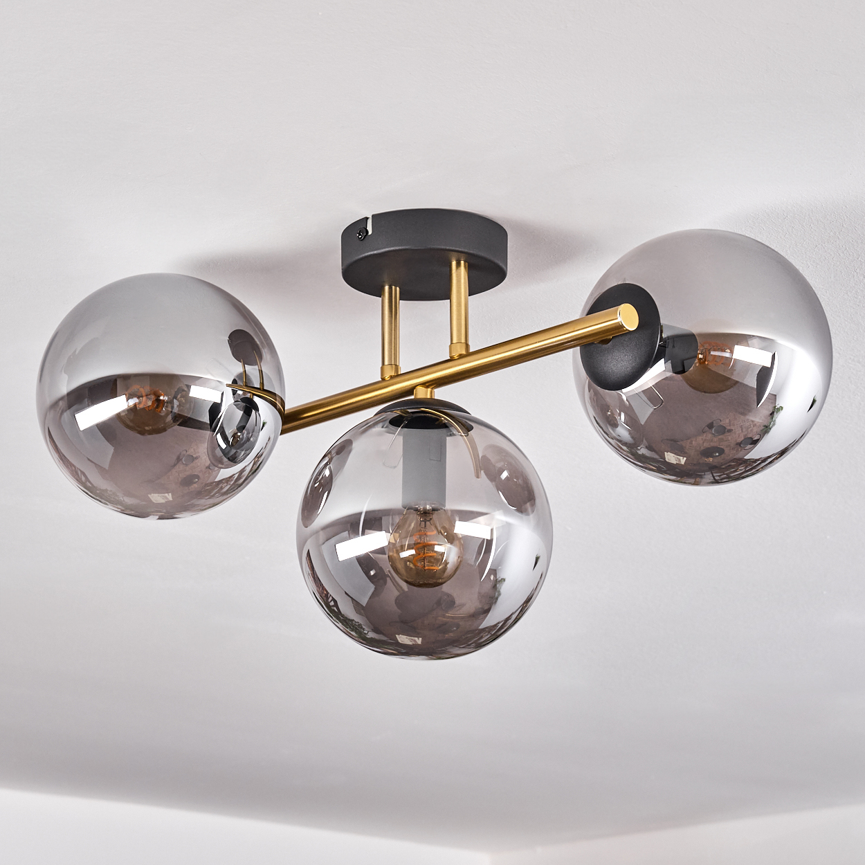 Eine moderne Deckenleuchte mit drei rauchigen Glaskugelschirmen. Die Leuchte hat goldene und schwarze Metallkomponenten und ist an einer weißen Decke montiert.