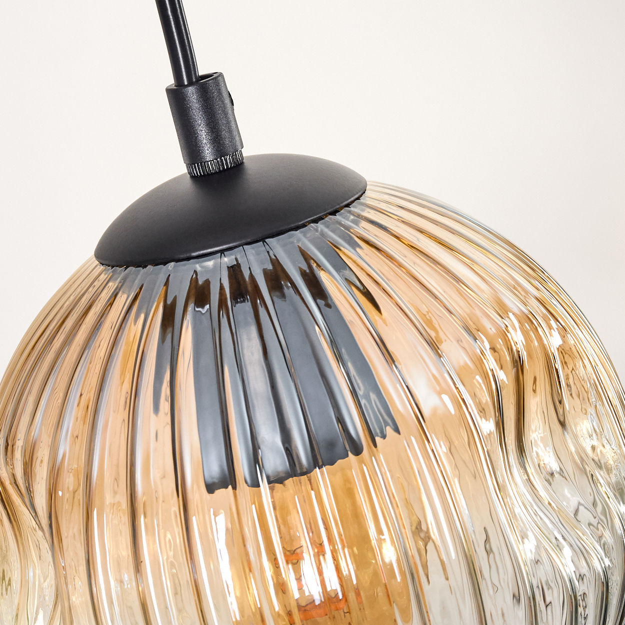 Nahaufnahme einer Glas-Pendelleuchte. Die Lampe hat einen wellenförmigen, bernsteinfarbenen Glasschirm und eine schwarze Metallhalterung.