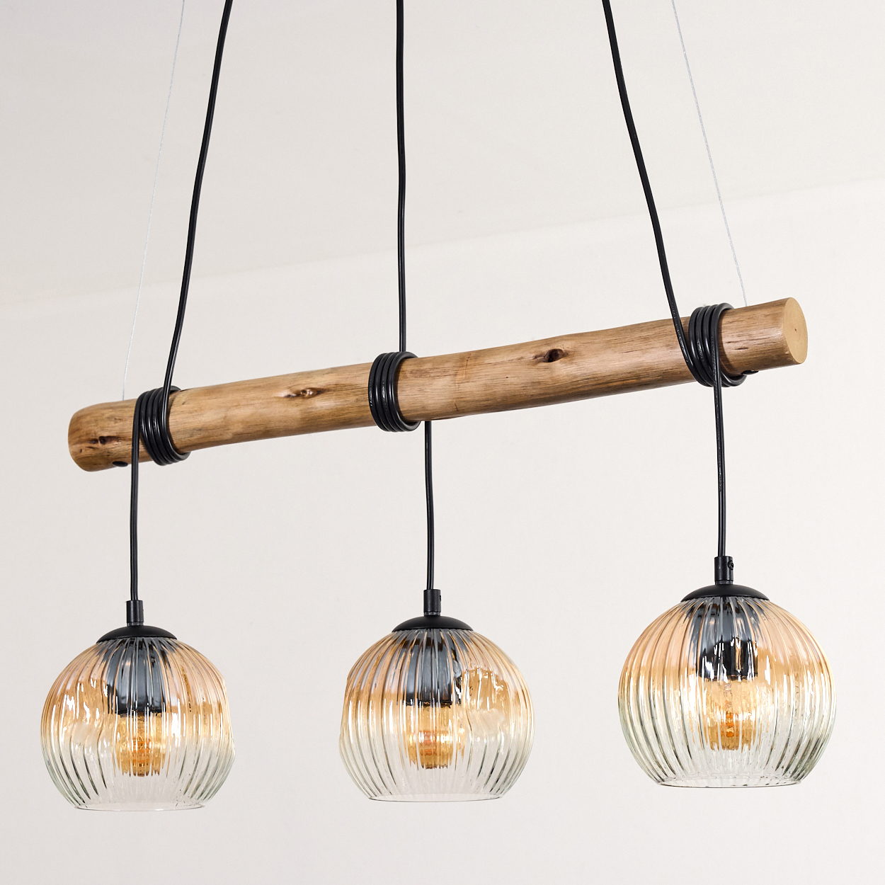 Ein Holzbalken mit drei hängenden Glaspendelleuchten, jede mit einer warmen Glühbirne. Schwarze Kabel und ein weißer Hintergrund.