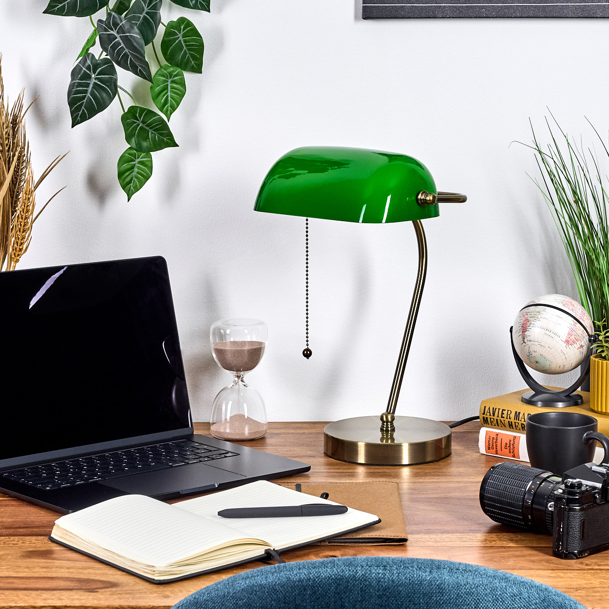 Ein Schreibtisch mit grüner Lampe, Laptop, Notizbuch, Kamera, Globus und Kaffeetasse. Pflanzen und eine Sanduhr sind ebenfalls vorhanden.