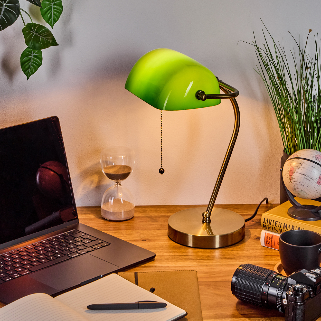 Ein Schreibtisch mit grüner Lampe, Laptop, Sanduhr, Kamera und Globus. Blätter an der Wand. Bücher und eine Kaffeetasse auf dem Schreibtisch.