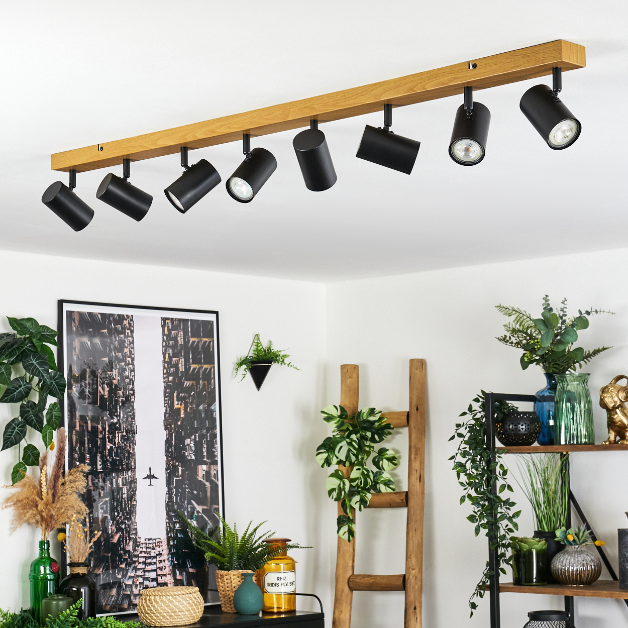 Modernes Interieur mit schwarzen Strahlern an einem Holzbalken. Pflanzen, Kunst und Deko auf Regalen und Tischen im Raum.