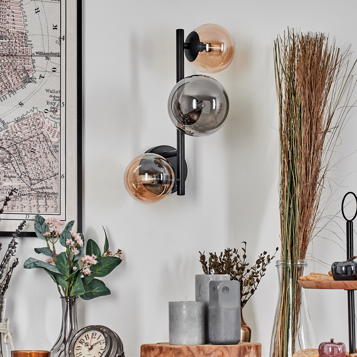 Eine Wandlampe mit drei Glaskugeln. Es gibt Vasen, Kerzen, eine Karte und dekorative Gegenstände. Der Hintergrund ist eine weiße Wand.