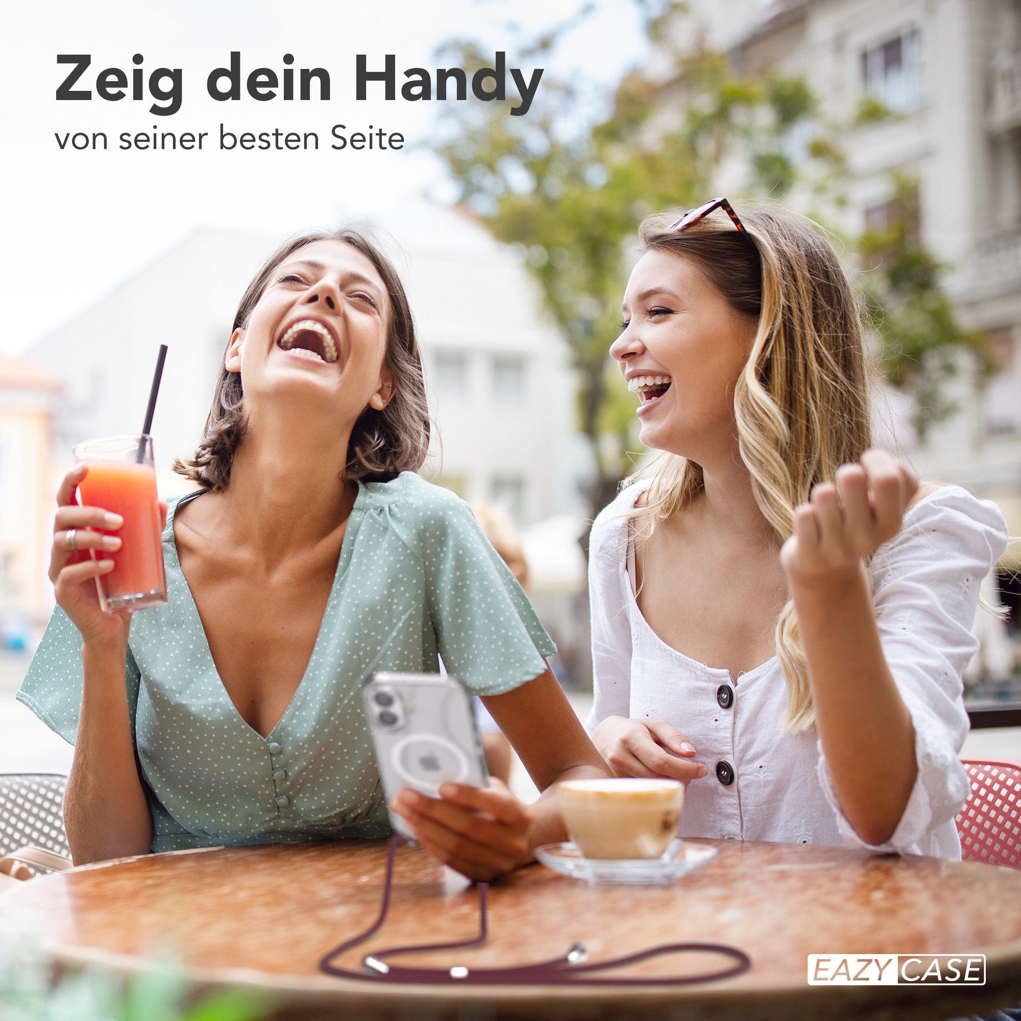 Zwei lachende Frauen in einem Café. Eine hält ein Handy, die andere ein Getränk. Tisch mit Kaffee.