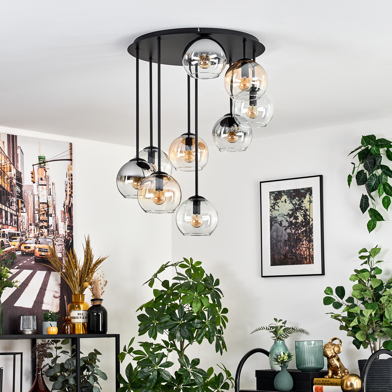 Ein moderner Kronleuchter mit Glaskugeln hängt in einem Raum mit Pflanzen, Kunstwerken und einem schwarzen Regal. Die Decke ist weiß.
