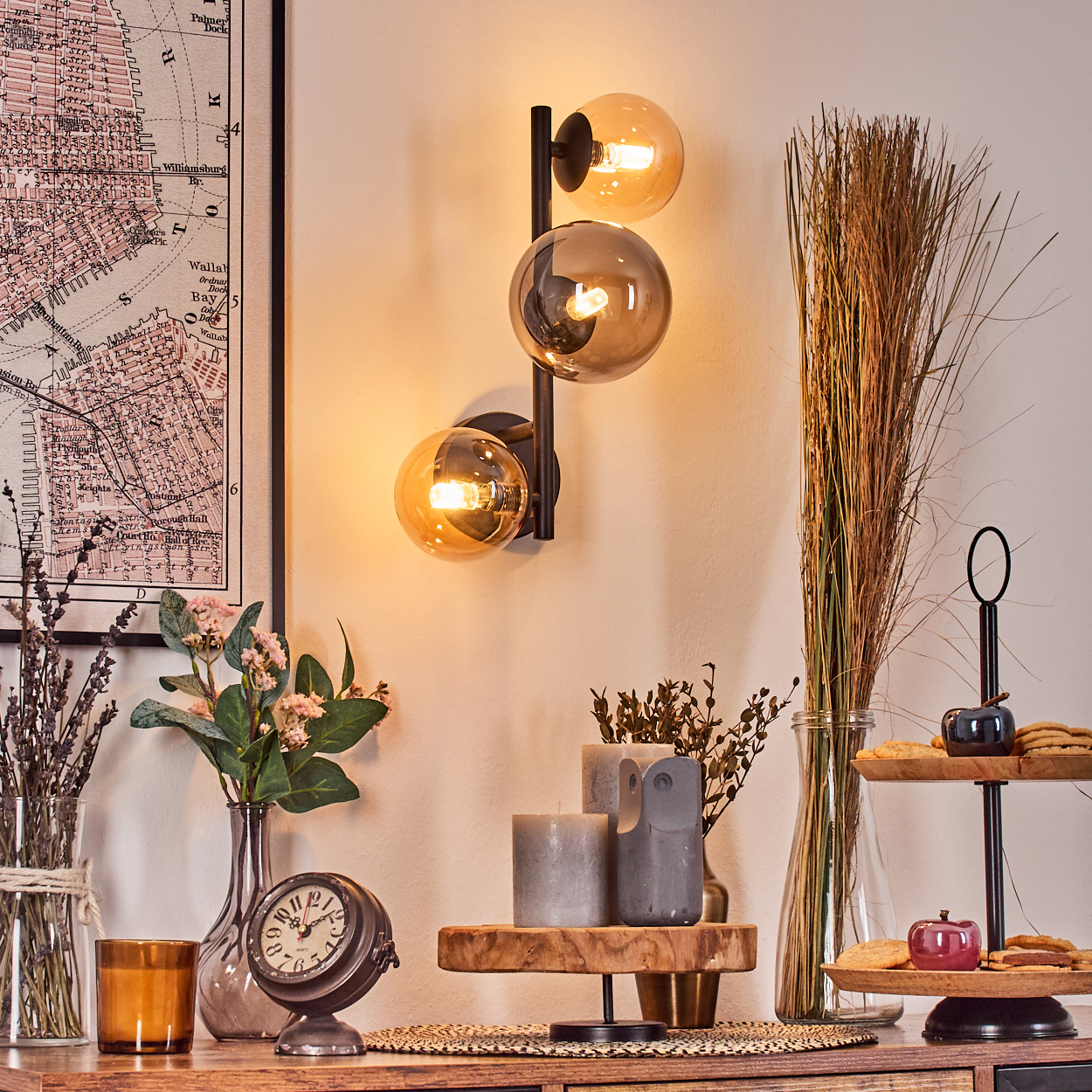 Eine dekorative Szene mit Wandlampe, Blumen, Uhr, Kerzen und Keksen auf einem Holztisch. Im Hintergrund ist eine Karte zu sehen.
