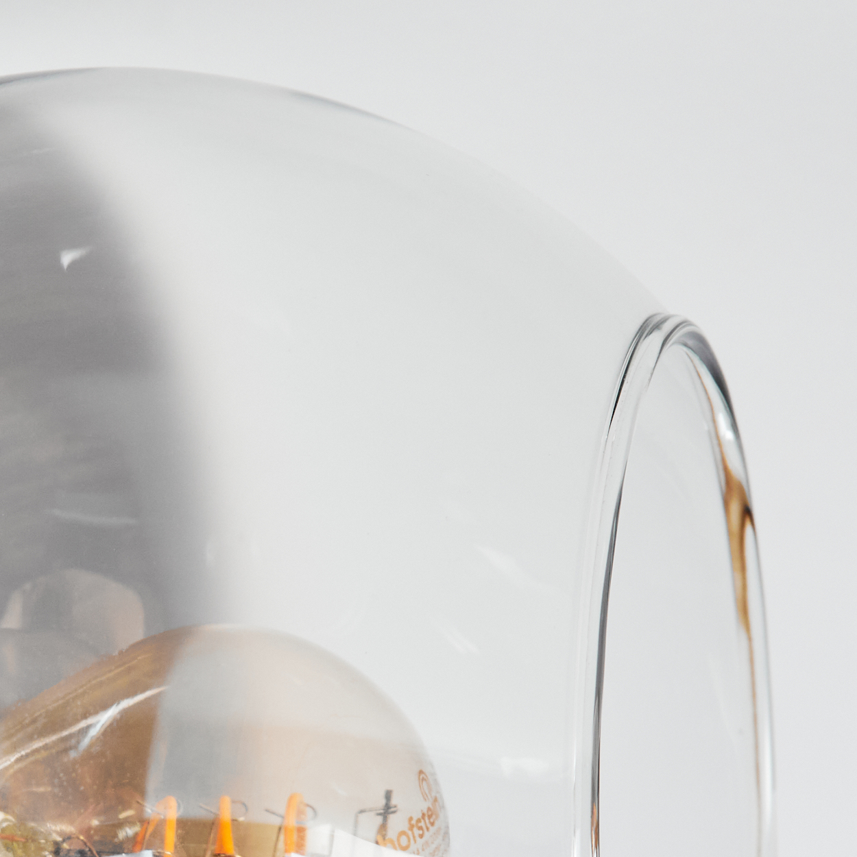 Nahaufnahme eines durchsichtigen Glasschirms mit einer Glühbirne. Der Hintergrund ist weiß. Der Schirm hat ein gebogenes Design.