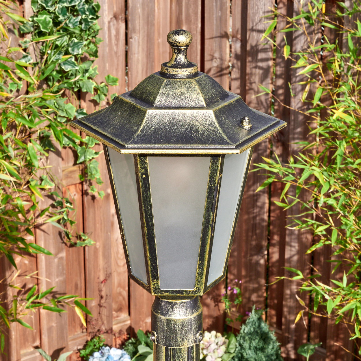 Eine verzierte Gartenlampe mit goldenem Oberteil und Rahmen, vor einem Holzzaun, umgeben von grünen Pflanzen.