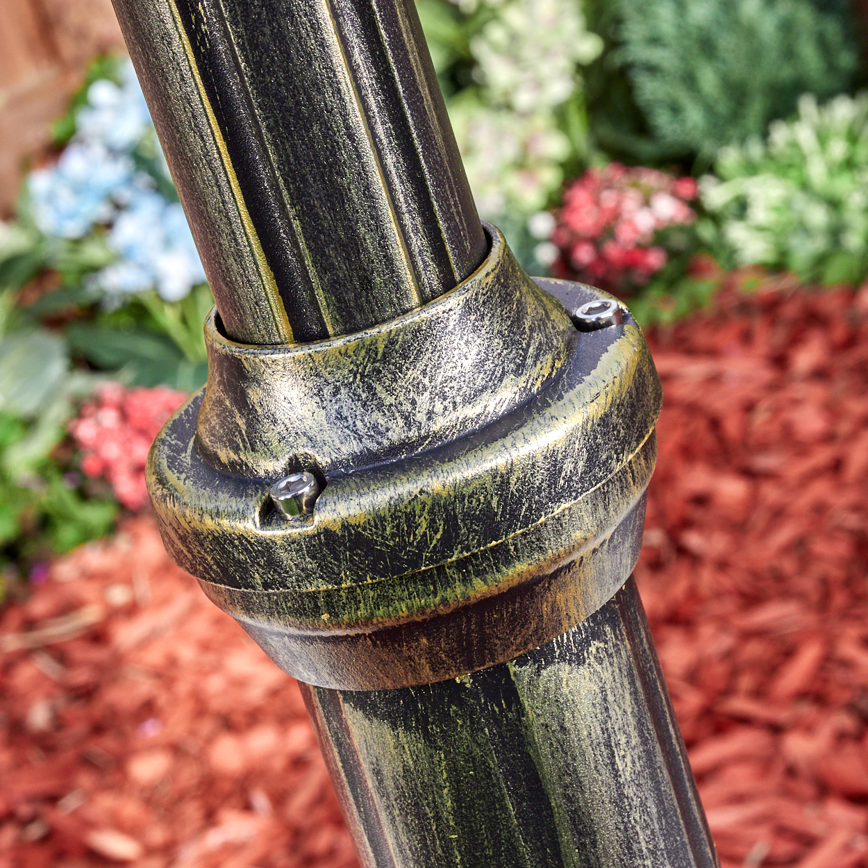 Nahaufnahme einer bronzefarbenen Metallstange mit Verbindungsring. Der Hintergrund zeigt Mulch, Blumen und Grün.