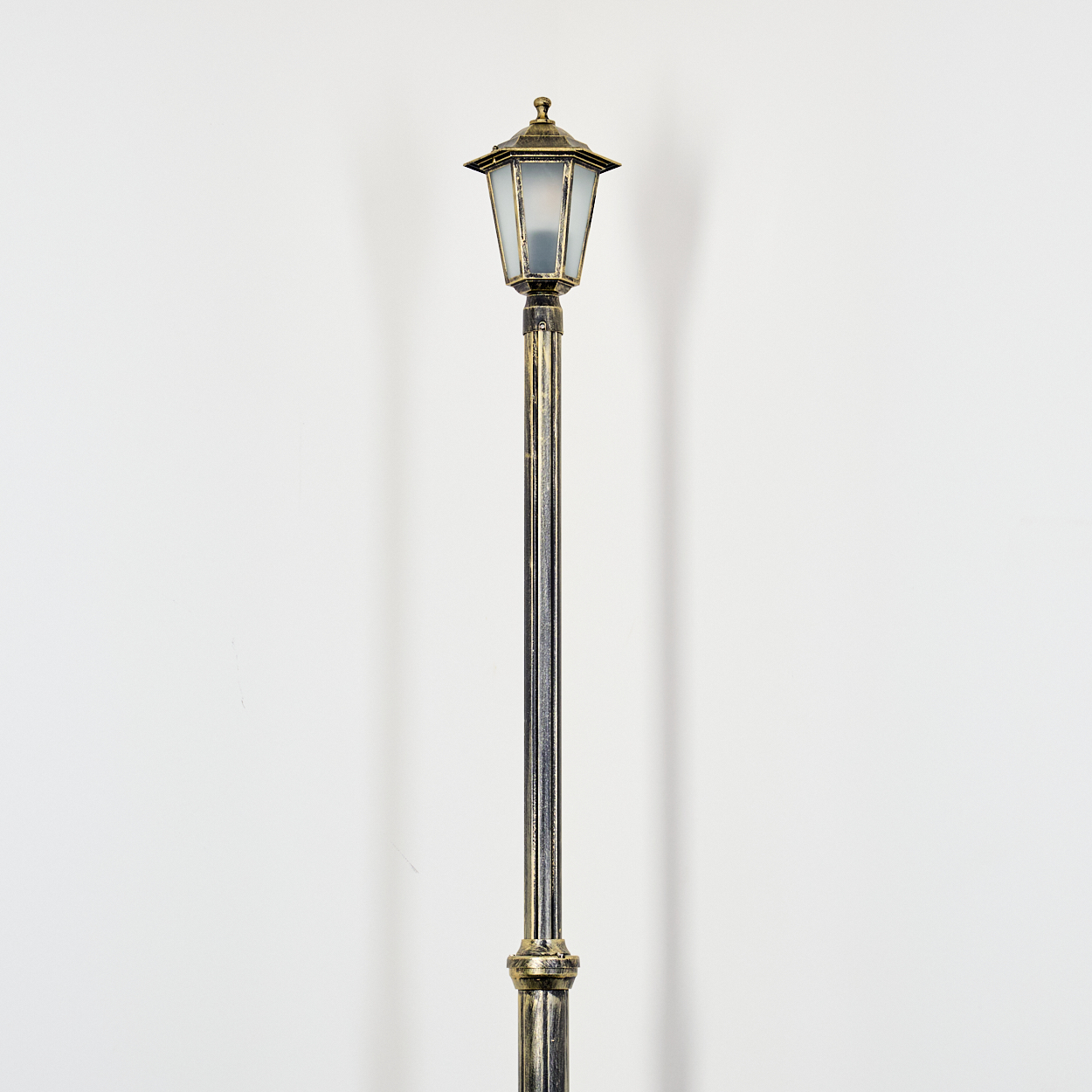 Eine dekorative Laterne mit bronzefarbenem Oberteil und Sockel steht vor einer weißen Wand. Die Lampe hat eine Glasscheibe.