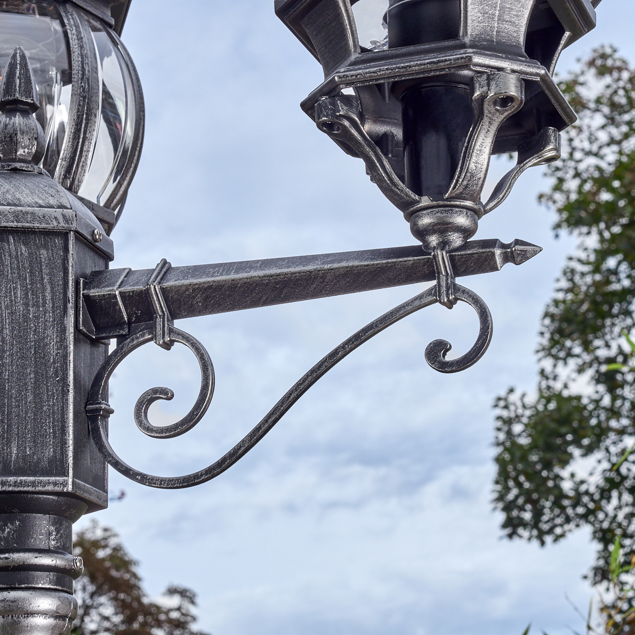 Nahaufnahme einer dekorativen Straßenlaterne. Die Lampe ist verziert, mit metallischem Finish, vor bewölktem Himmel und grünem Laub.