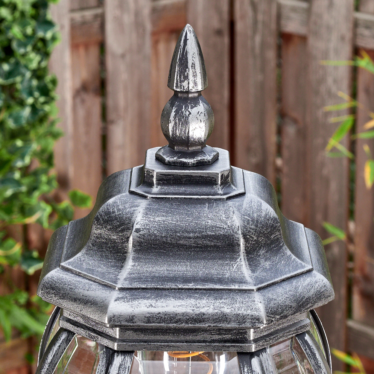 Nahaufnahme einer dekorativen Außenlampe mit silberfarbener Oberseite und Holzzaun im Hintergrund.