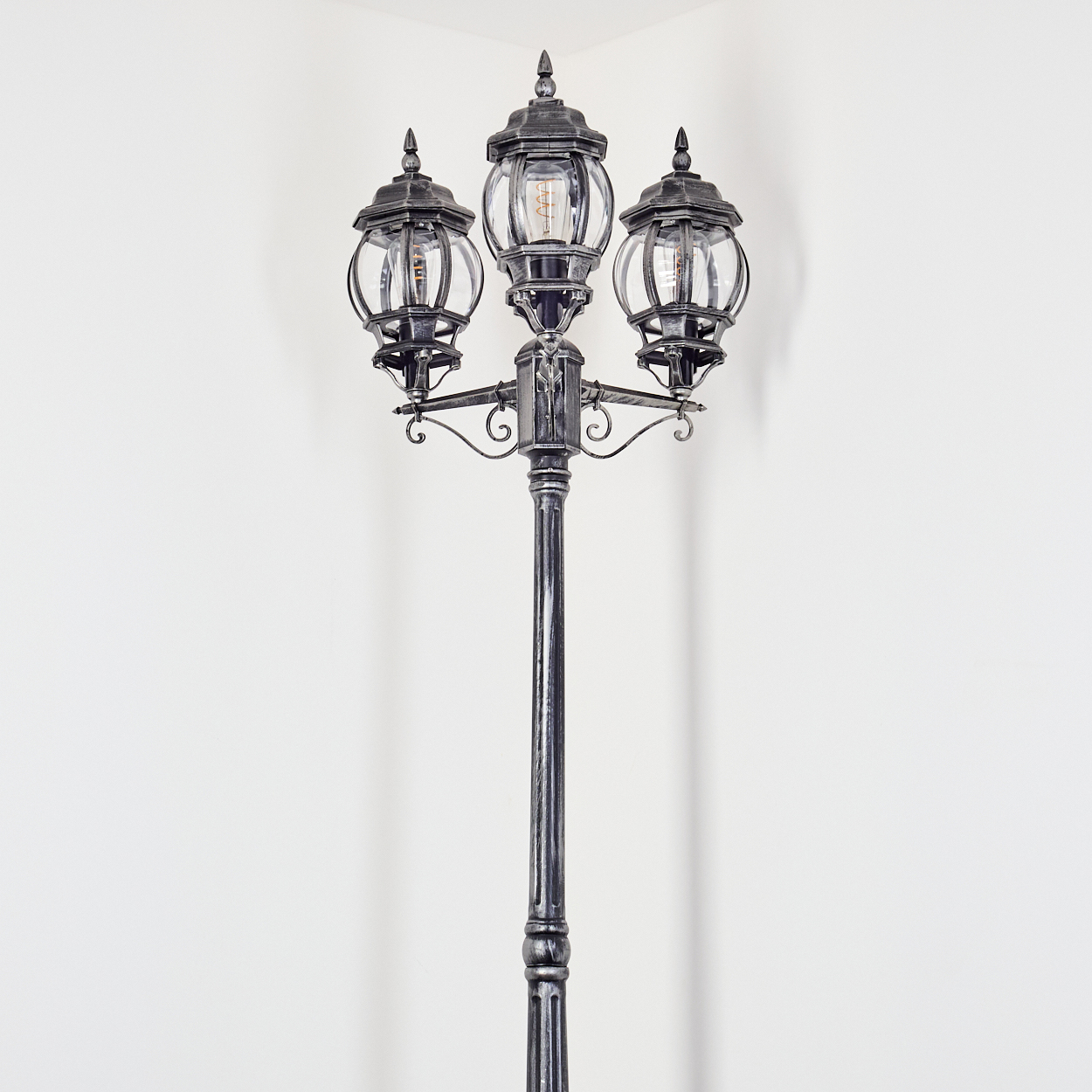 Eine dekorative Laterne mit drei Lampen. Die Lampen sind in Glas eingeschlossen. Der Pfosten ist schwarz und steht vor einer weißen Wand.