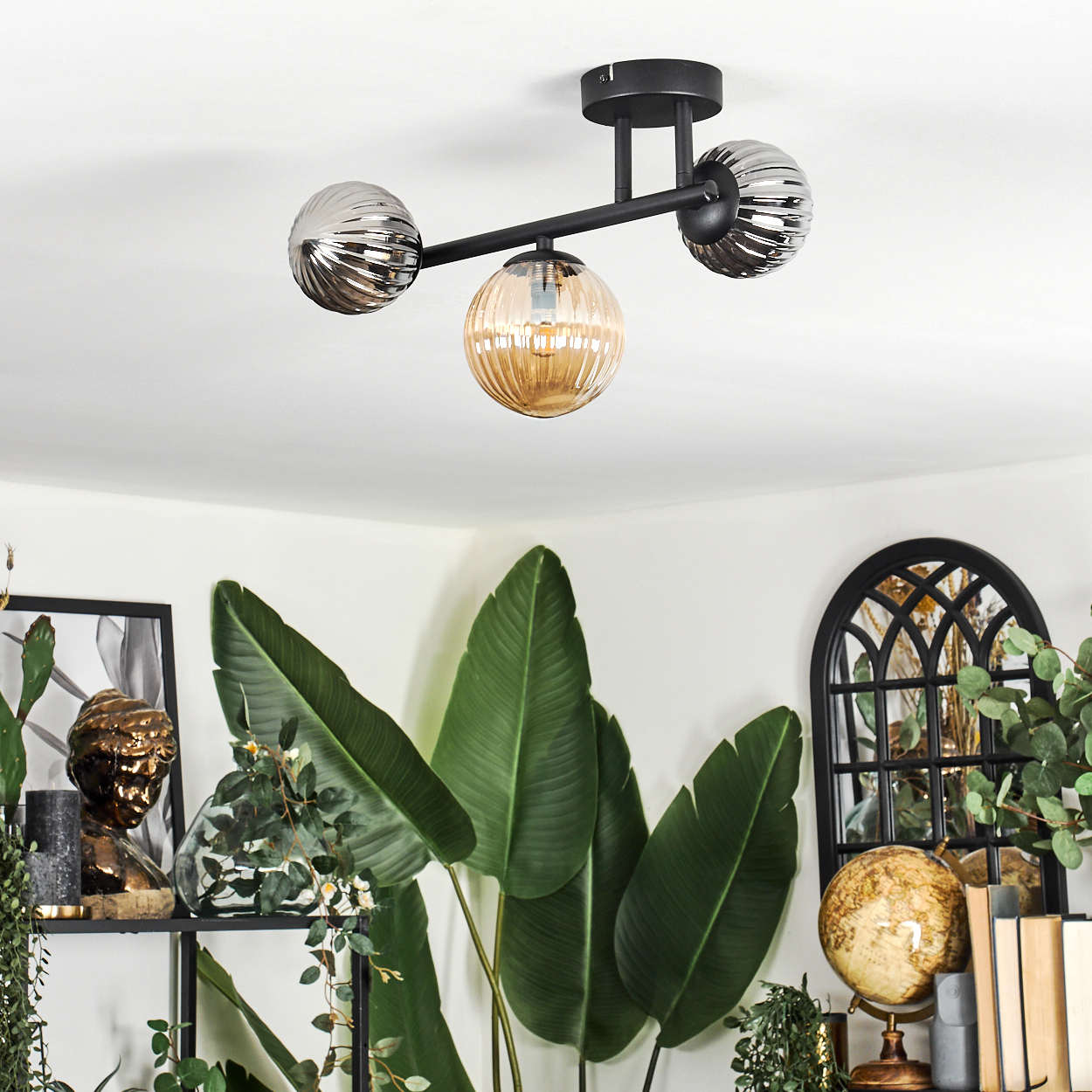 Eine moderne Deckenlampe mit drei Glaskugeln hängt über großen grünen Blättern. Dekorative Gegenstände, darunter ein Globus und Bücher, sind sichtbar.