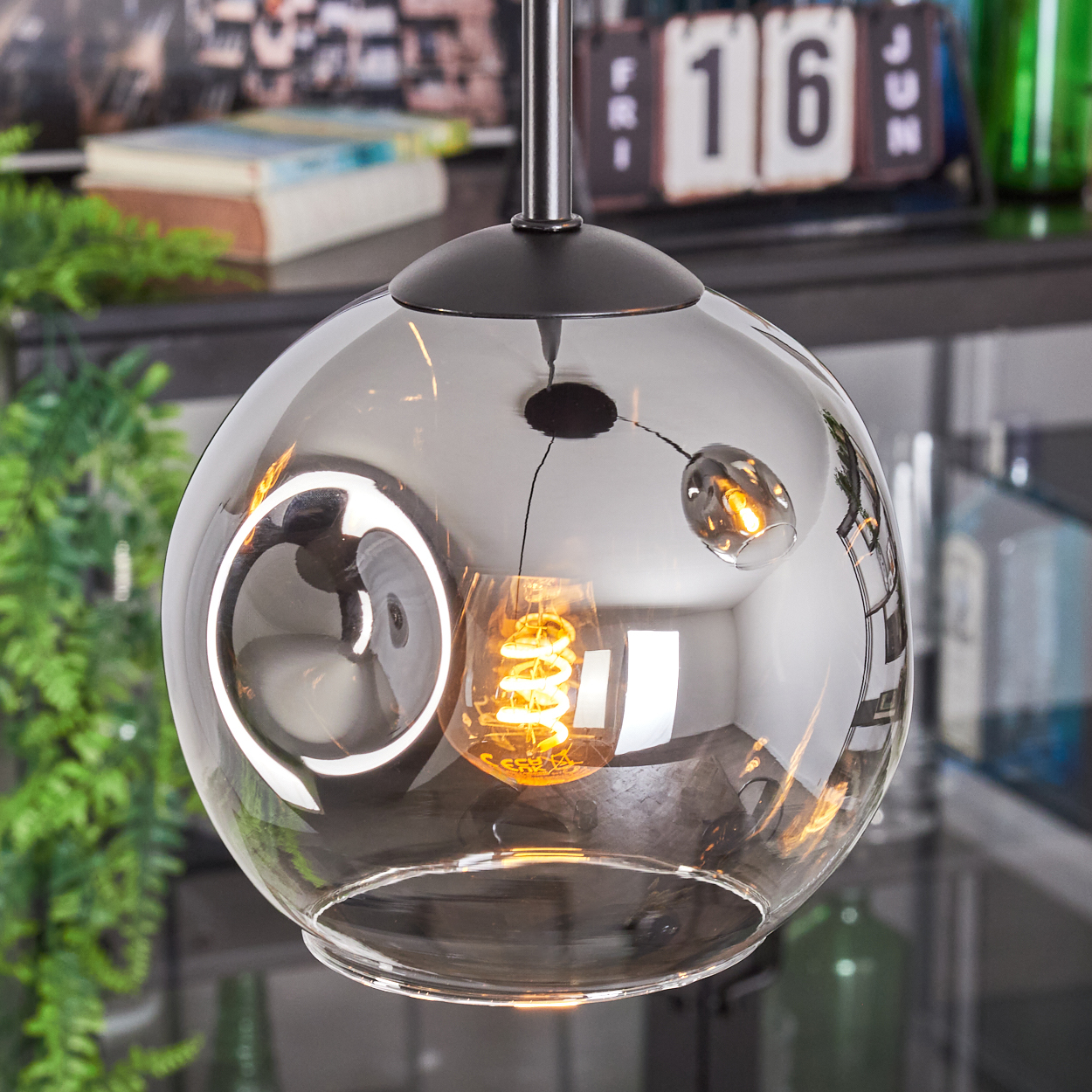 Eine moderne Glaskugel-Pendelleuchte mit einer Wendelglühbirne. Der Hintergrund zeigt einen Kalender, Grün und andere Objekte.