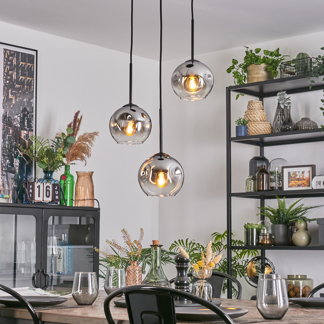 Esszimmer mit Hängelampen, gedecktem Tisch. Regale mit Pflanzen und Dekoration. Schwarze Stühle. Glasvasen mit Blumen.
