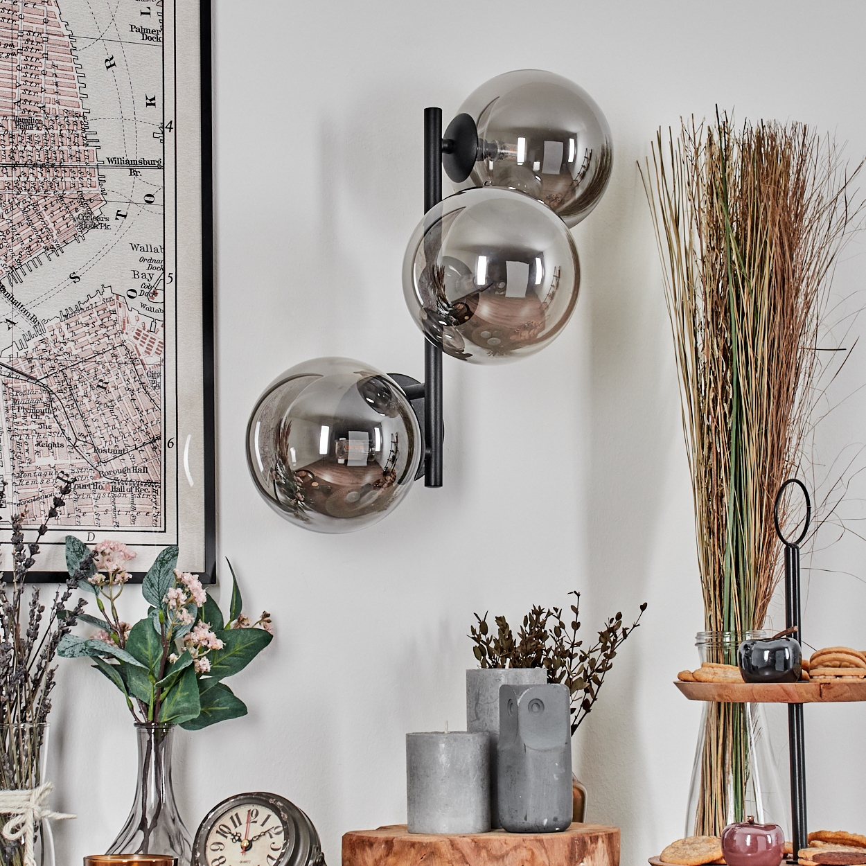 Eine Wand mit einer Karte, einer Lampe mit drei Kugeln, Blumen und dekorativen Gegenständen. Die Lampe hat graue Kugeln. Es gibt Kerzen und eine Uhr.
