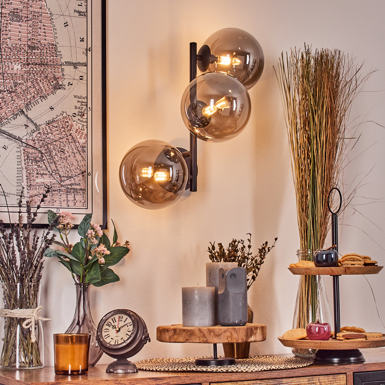 Eine Zimmerecke mit Lampe, Karte, Blumen, Uhr, Kerzen und einem mehrstöckigen Tablett mit Keksen. Die Wand ist beige.