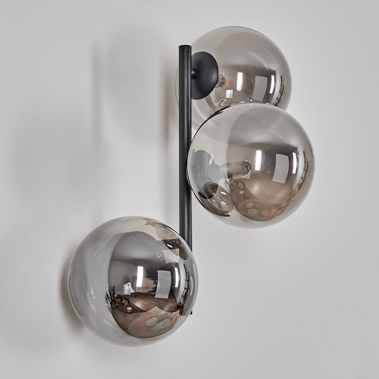 Moderne Wandlampe mit drei rauchfarbenen Glaskugeln. Schwarzer Metallrahmen. Spiegelung des Raumes in den Kugeln. An weißer Wand montiert.
