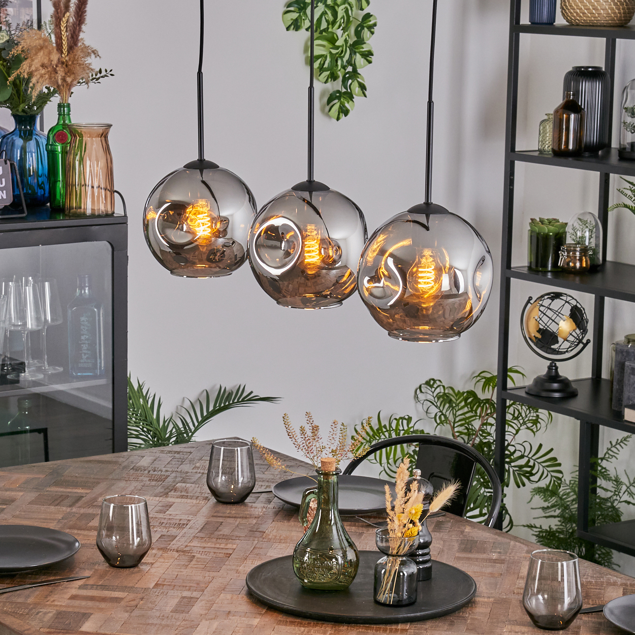 Esszimmer mit Holztisch, drei Hängelampen und verschiedenen Dekorationsartikeln. Pflanzen und Gläser stehen auf dem Tisch.