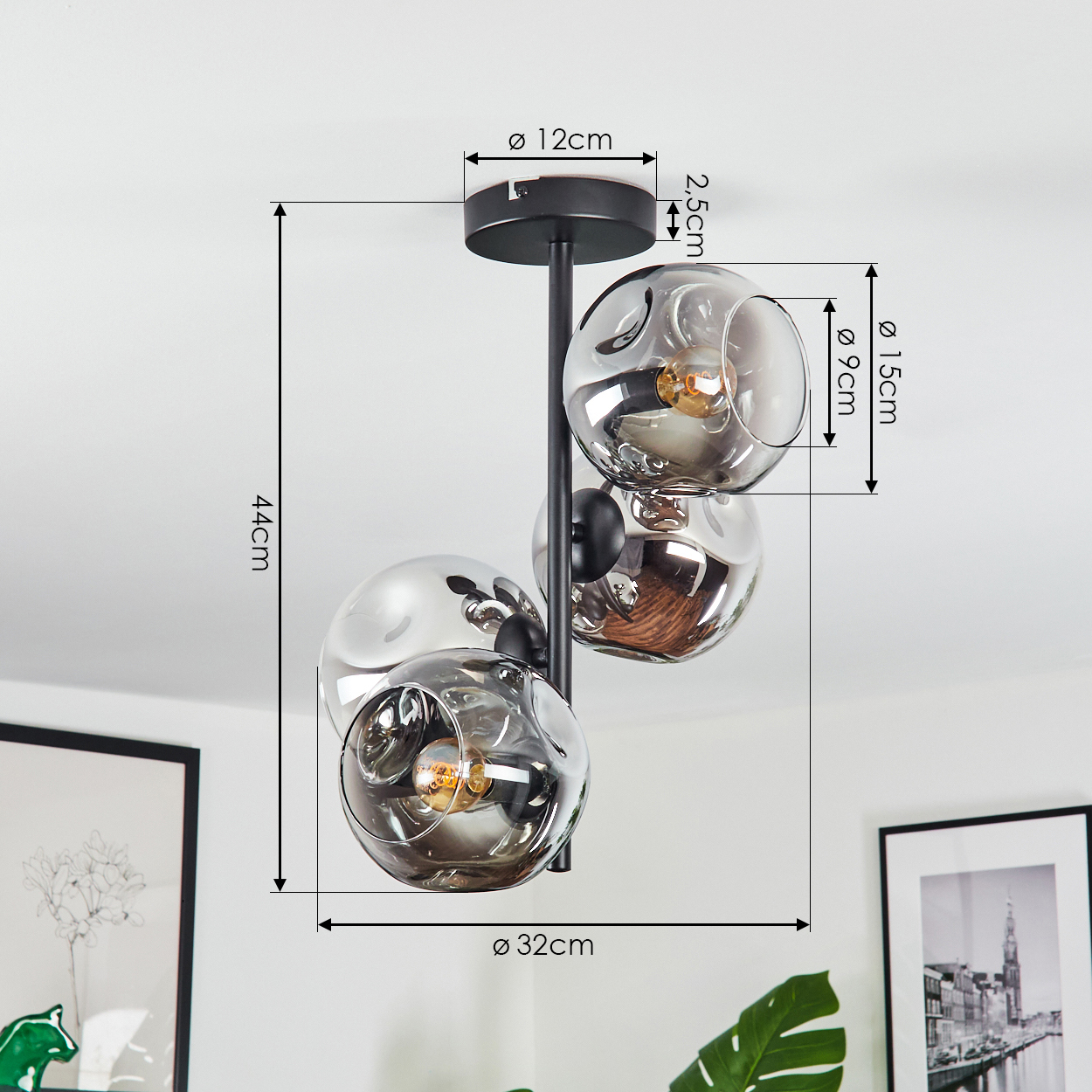 Eine moderne Deckenlampe mit drei kugelförmigen Glasschirmen. Schwarzer Rahmen, Maße sind angegeben. Der Hintergrund zeigt gerahmte Kunst und eine Pflanze.