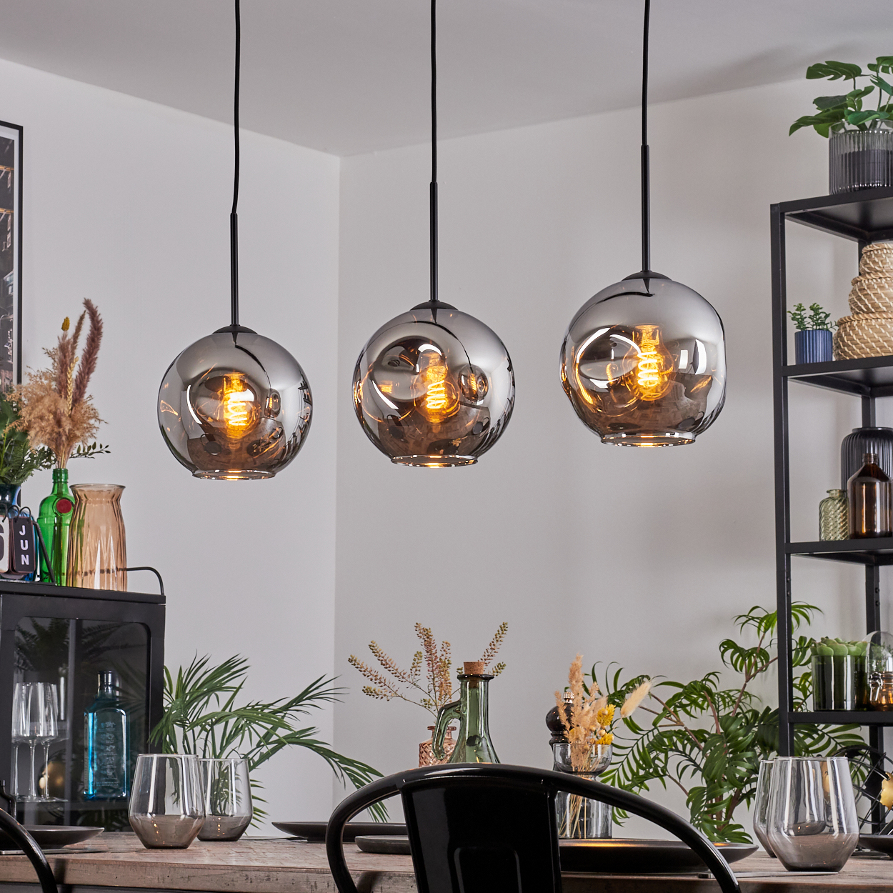Drei Pendelleuchten mit rauchfarbenen Glasschirmen hängen über einem Tisch. Die Szene umfasst Pflanzen, Gläser und ein schwarzes Metallregal.