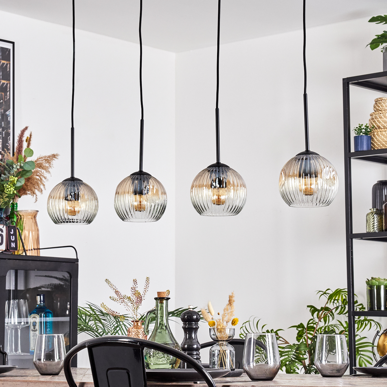 Vier Glas-Pendelleuchten hängen über einem Esstisch mit Gläsern und Dekorationen. Pflanzen und Regale sind im Hintergrund zu sehen.