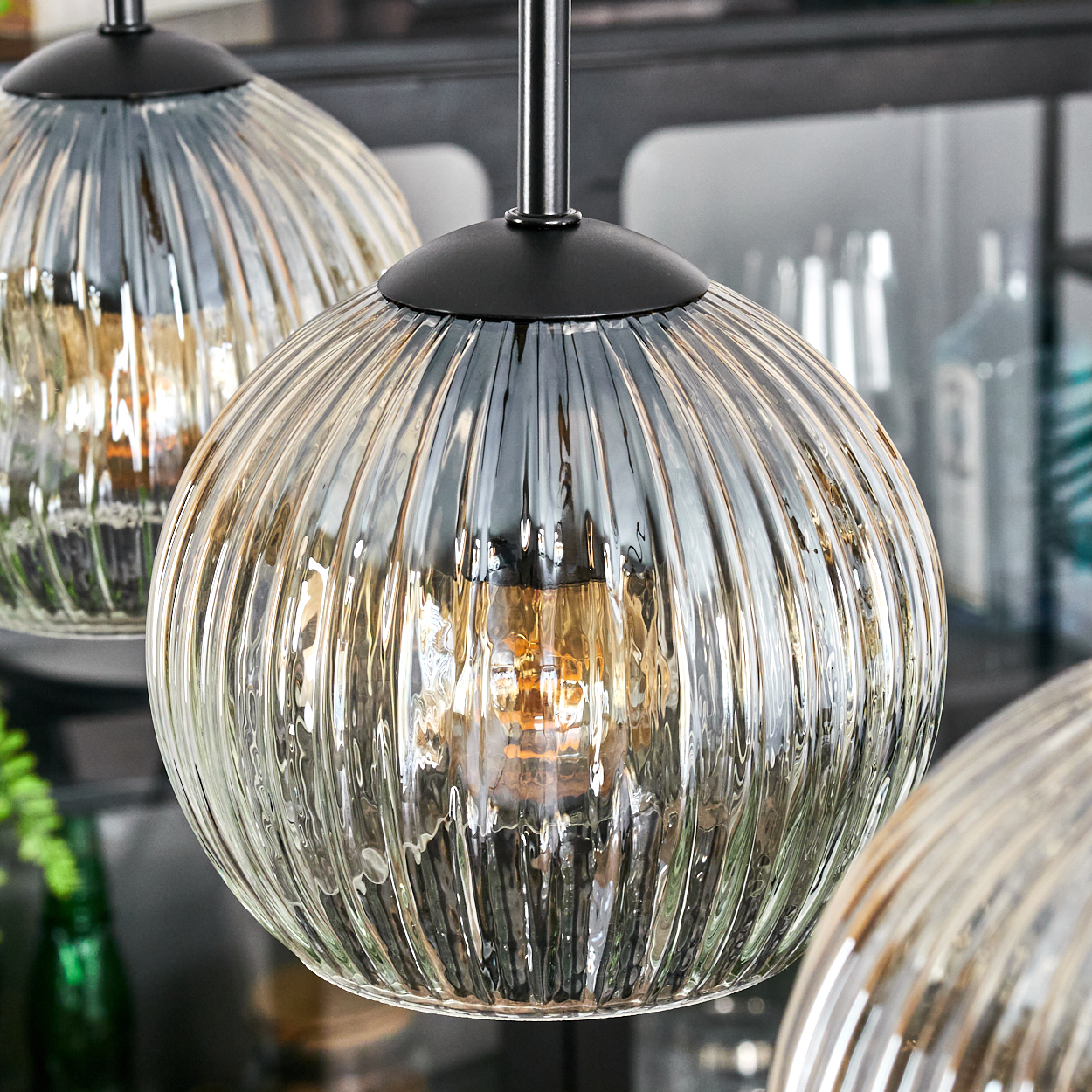 Drei Pendelleuchten aus Glas mit geripptem Design. Schwarze Metallbefestigungen. Im Hintergrund sind verschwommene Regale und Flaschen zu sehen.