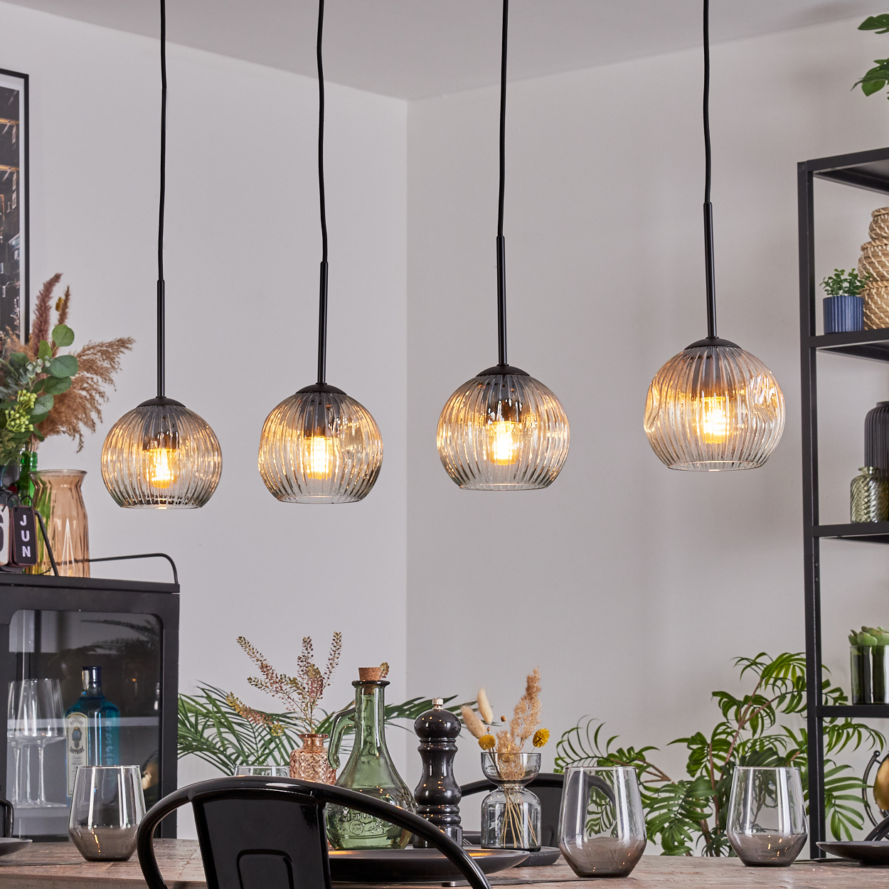 Vier Glaskugel-Pendelleuchten hängen über einem Tisch mit Gläsern und Pflanzen. Der Hintergrund zeigt ein Regal und andere Dekorationen.