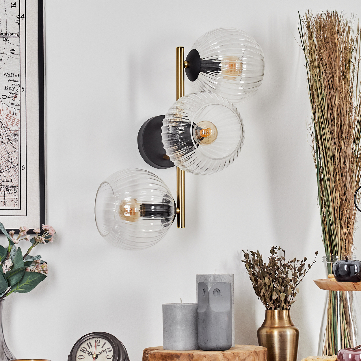 Eine Wandlampe mit drei Glaskugeln. Es gibt Kerzen, eine Uhr, getrocknete Pflanzen und eine Karte an der Wand.