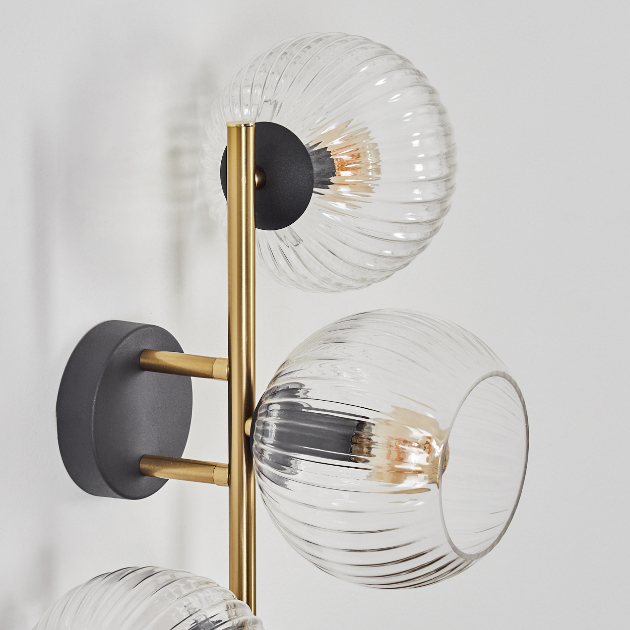 Eine moderne Wandlampe mit drei klaren, geriffelten Glaskugelschirmen. Getragen von Messingarmen und einer schwarzen Mittelstütze an einer weißen Wand.