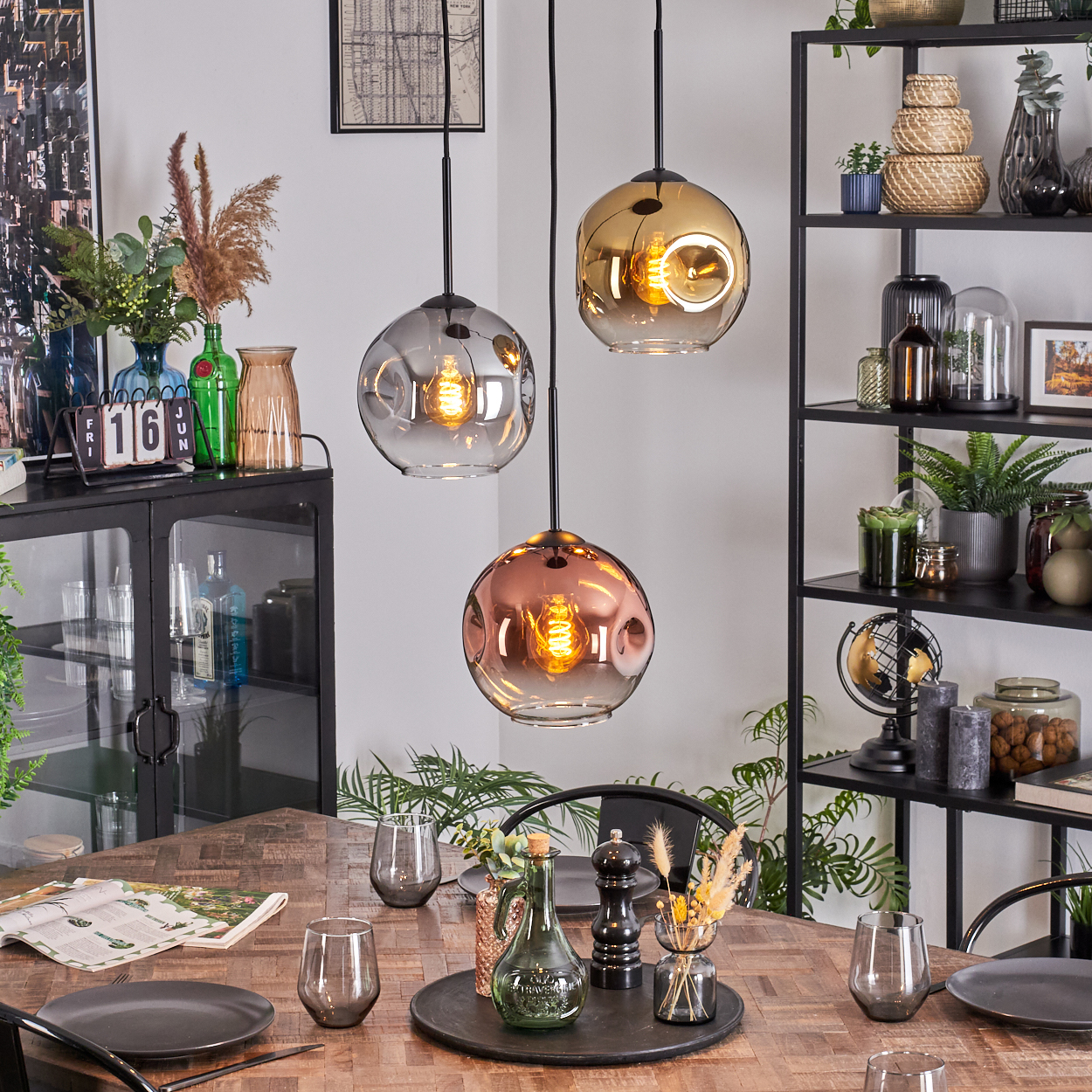 Esszimmer mit Holztisch, drei Pendelleuchten und Dekorationsartikeln. Schwarze Regale mit Pflanzen und Ornamenten.