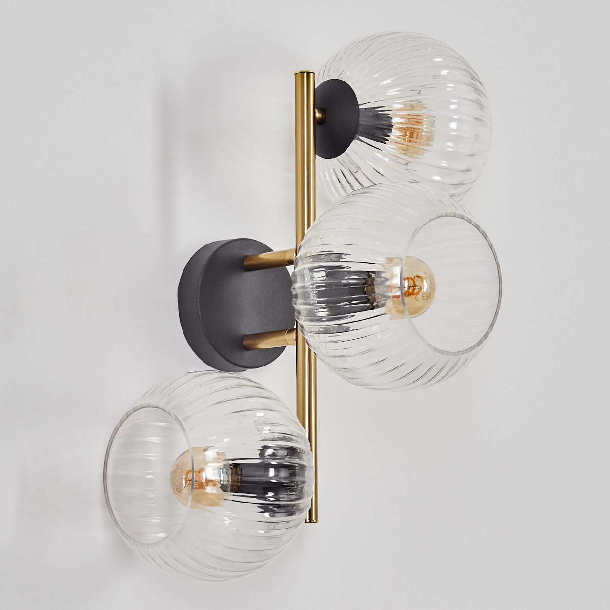 Moderne Wandlampe mit drei Glaskugeln. Sie hat einen goldenen Rahmen und einen schwarzen Sockel, montiert an einer weißen Wand.