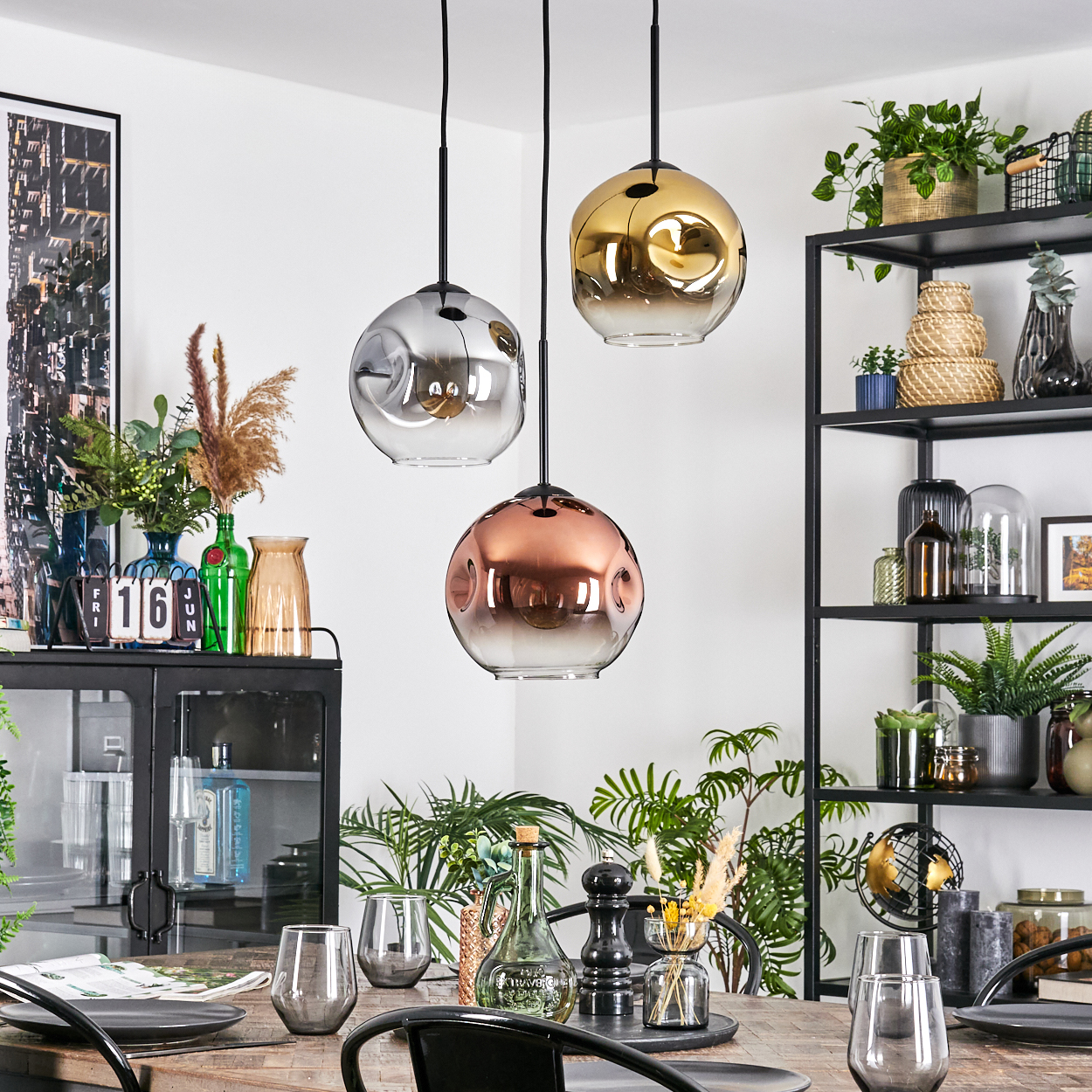Esszimmer mit drei Hängelampen. Tisch mit Gläsern und Dekor. Regale mit Pflanzen und Dekor im Hintergrund.