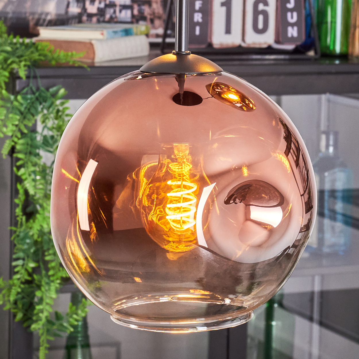Eine kupferfarbene Glas-Pendelleuchte mit einer Wendelglühbirne. Im Hintergrund sind Blätter und ein Kalender zu sehen.