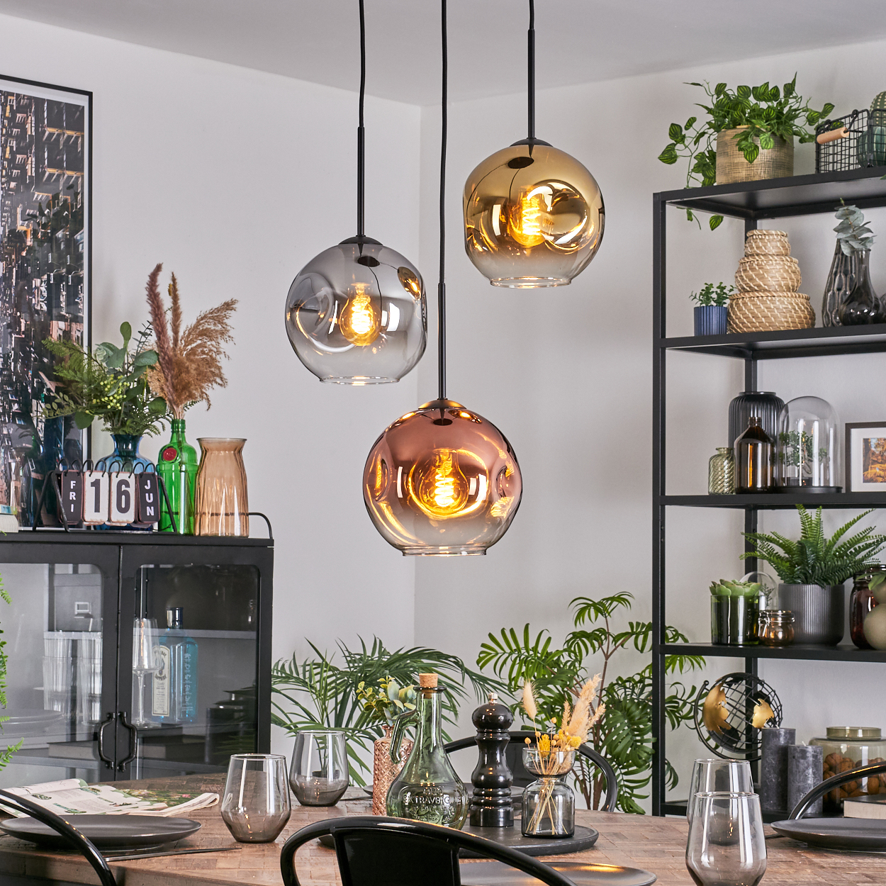 Drei Pendelleuchten über einem Holztisch mit Gläsern und Tellern. Regale mit Pflanzen und Dekoration sind im Hintergrund sichtbar.