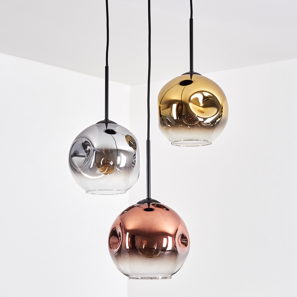 Drei kugelförmige Pendelleuchten, silber, gold und kupfer, hängen vor einem weißen Eckhintergrund.