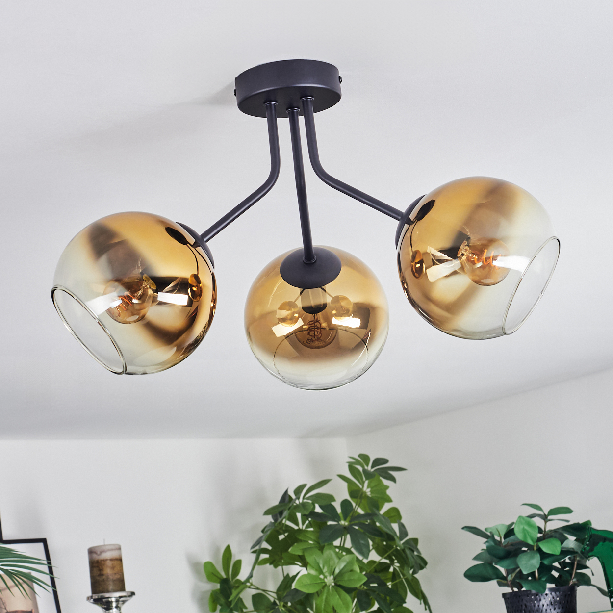 Eine moderne Deckenleuchte mit drei bernsteinfarbenen Glaskugeln. Schwarze Metallarme und Deckenhalterung. Grüne Pflanzen im Hintergrund.